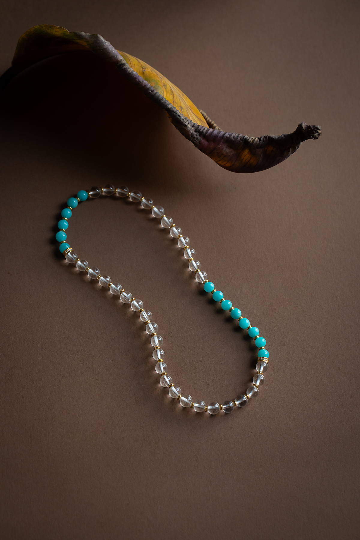 A strand of clear quartz and turquoise beads curved on brown paper beneath a dried leaf.