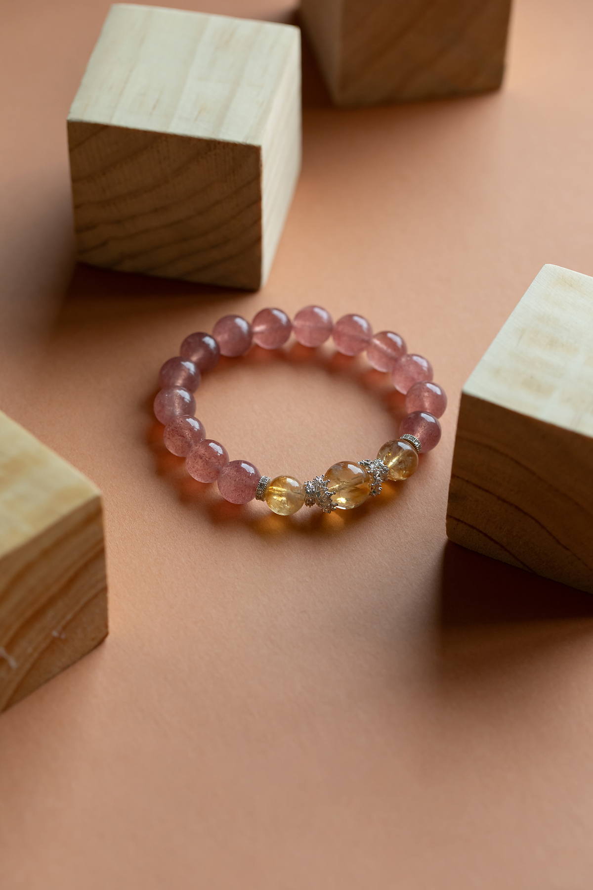 A custom crystal bracelet of strawberry quartz and citrine beads standing among pale wooden blocks.